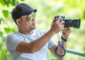 Man in white shirt using a camera outdoors, surrounded by greenery.